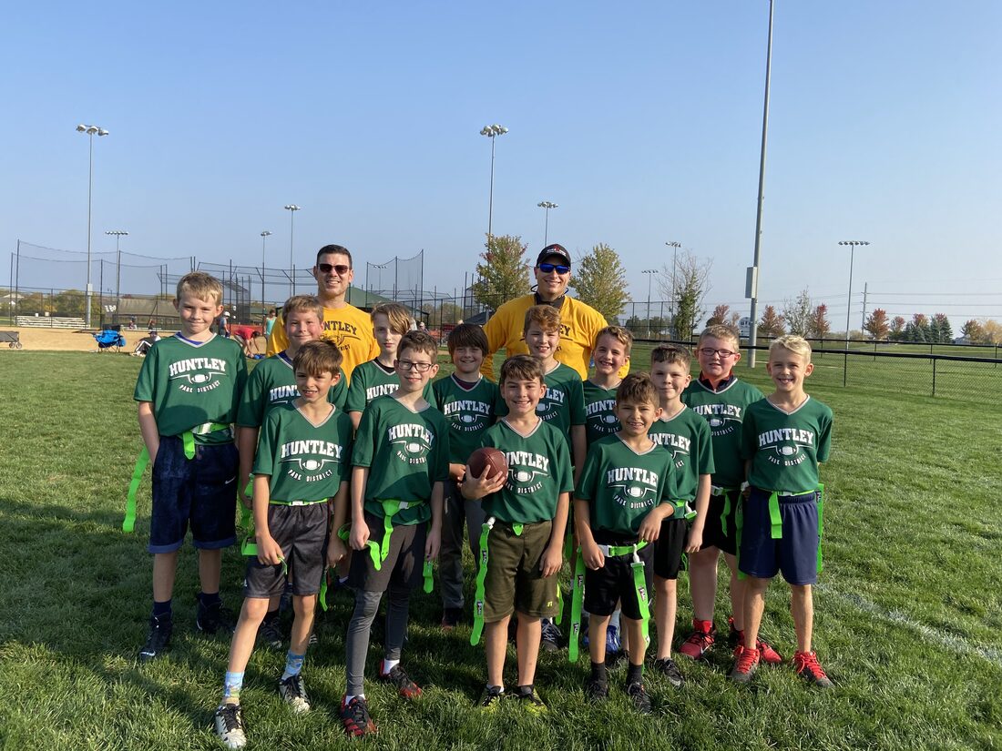 A Huntley youth flag football team in green jerseys lined up on the field with their coaches, including Aaron Holbrook, under floodlights at a Huntley, Illinois sports park.