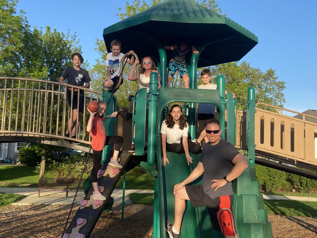 Aaron Holbrook sitting on a playground slide in Huntley, Illinois, surrounded by children of the summer park group climbing over the green play structure above and around him, one holding a basketball.