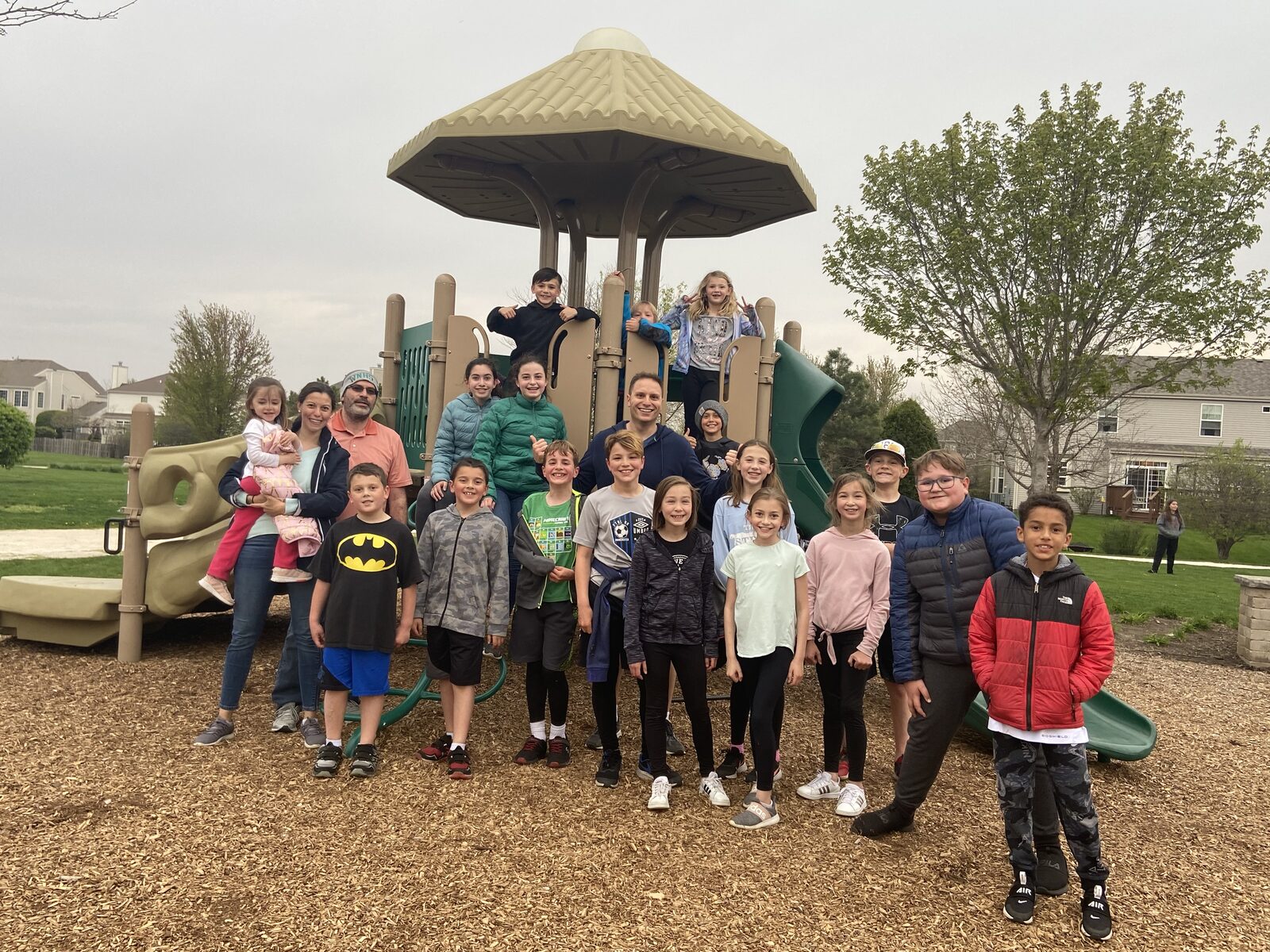 The Huntley summer park group organized by Aaron Holbrook, gathered on the playground at the end of an evening. About twenty children ranging from roughly five years old to high school age stand with several parents in front of a wooden play structure with a green slide, in a Huntley, Illinois neighborhood park backed by houses and open grass.