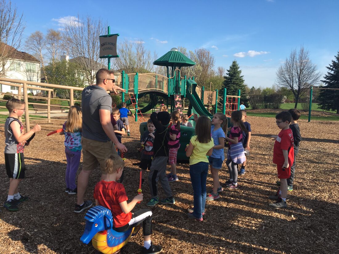 Aaron Holbrook standing with the children of his Huntley summer park group while they vote on which game to play next, a loose circle of kids around him on the wood chips beside the playground, with a younger child on a spring rocker in the foreground.